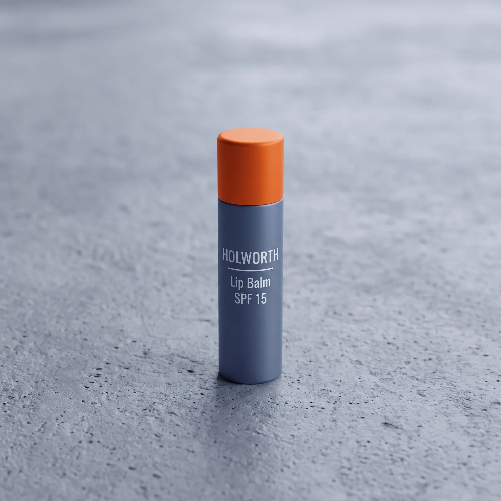 A steel-grey stick of Holworth Lip Balm SPF 15 with an orange cap, standing on grey concrete.