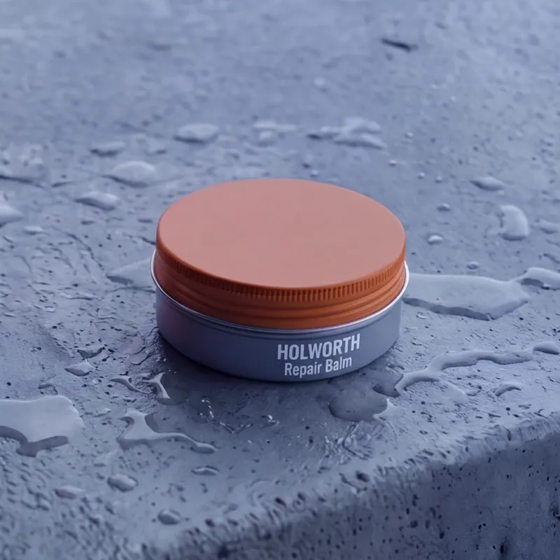 An open steel-grey tin of Repair Balm, its orange lid beside it, one finger swipe in the balm.
