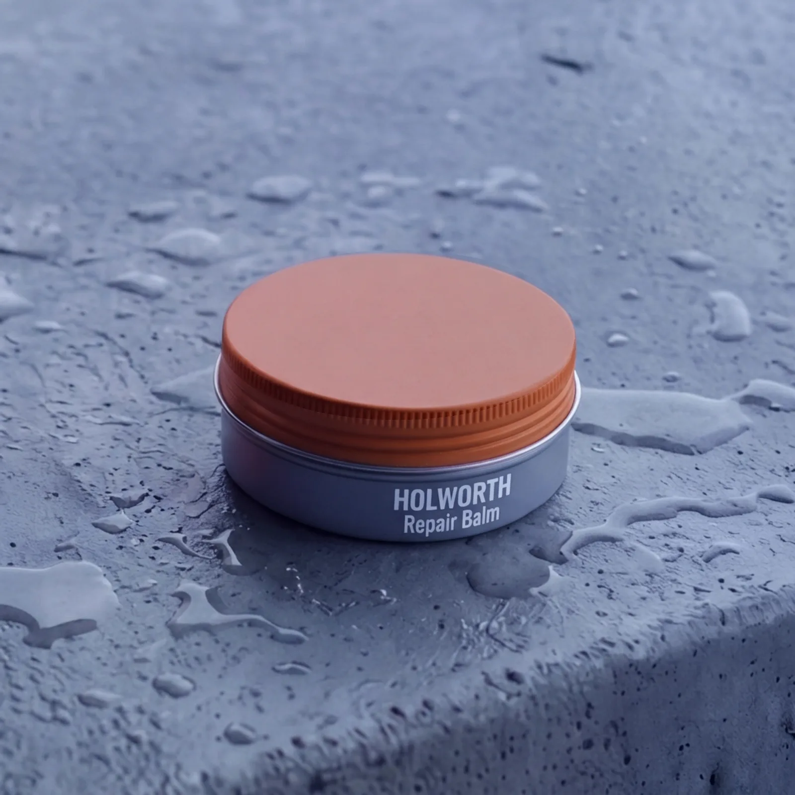 Holworth Repair Balm, a slate-grey container with an orange cap, labeled HOLWORTH, on grey concrete.