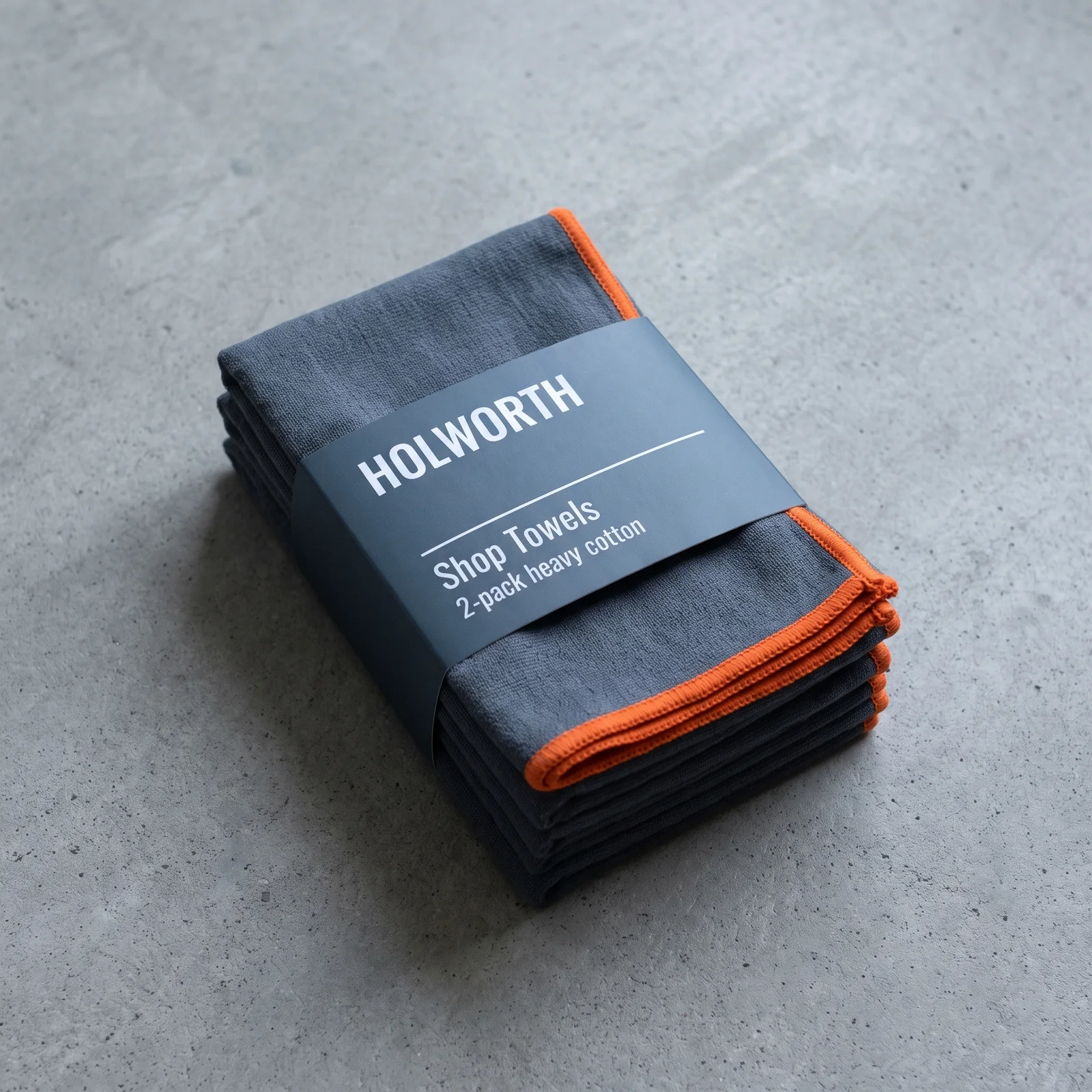 A stack of folded dark grey Holworth shop towels with orange edging, on grey concrete.